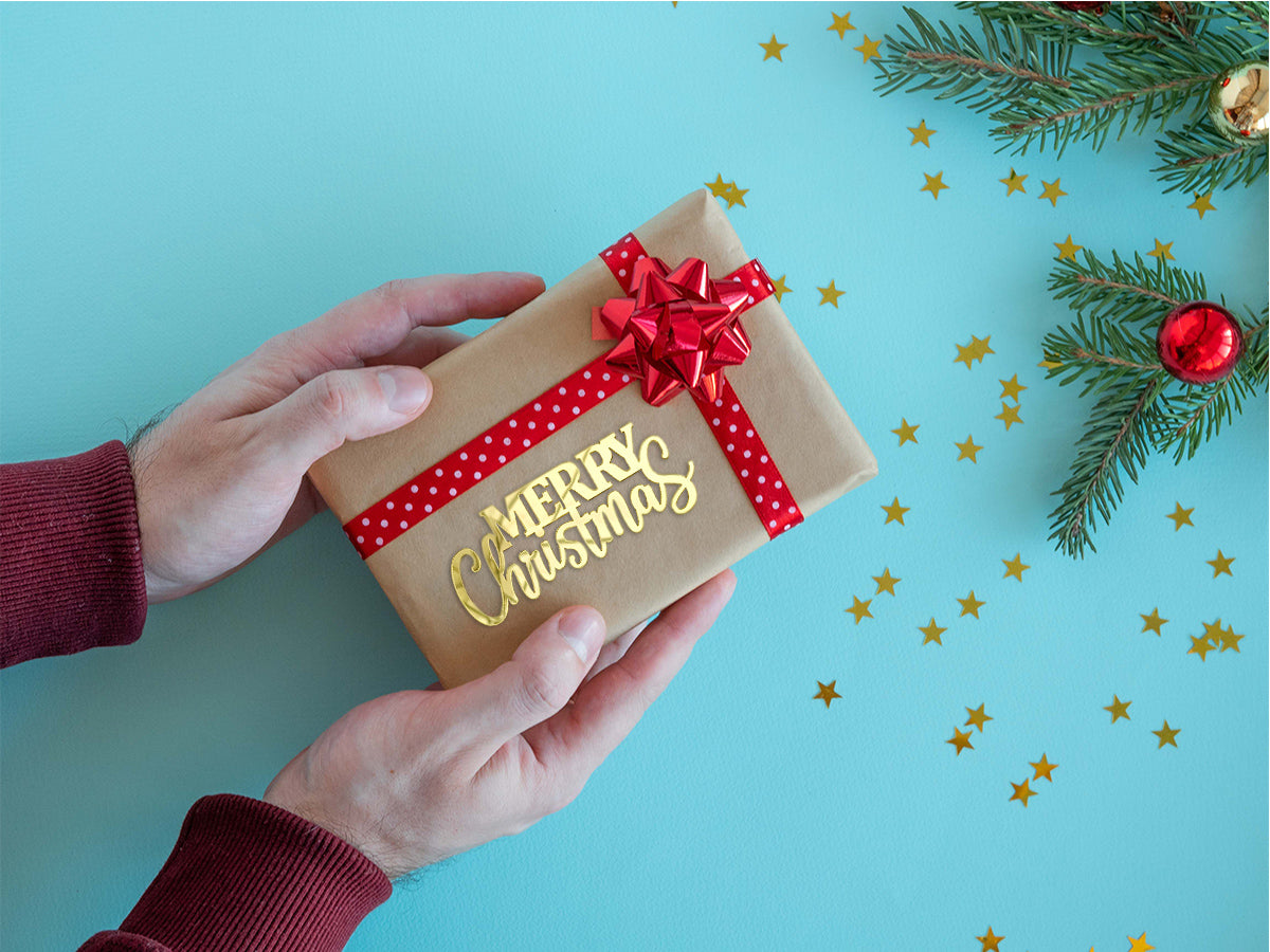 Person holding a Christmas gift wrapped in brown paper with a red bow on a blue background with gold stars and greenery.